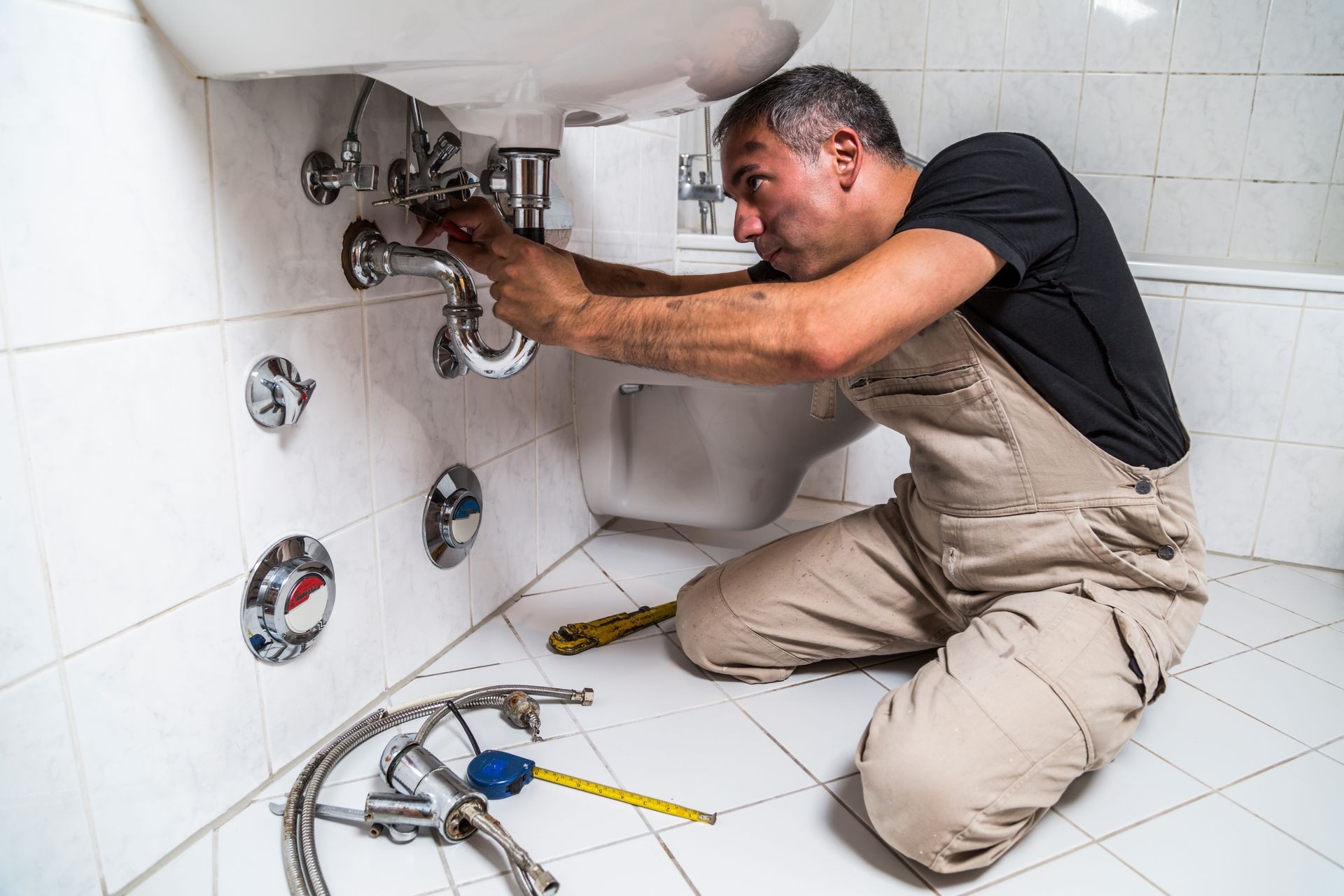 Specialist male plumber in overalls and black t-shirt repairs faucet with yellow old shabby screwdriver in bathroom on the floor near screwdriver, hammer, pliers
